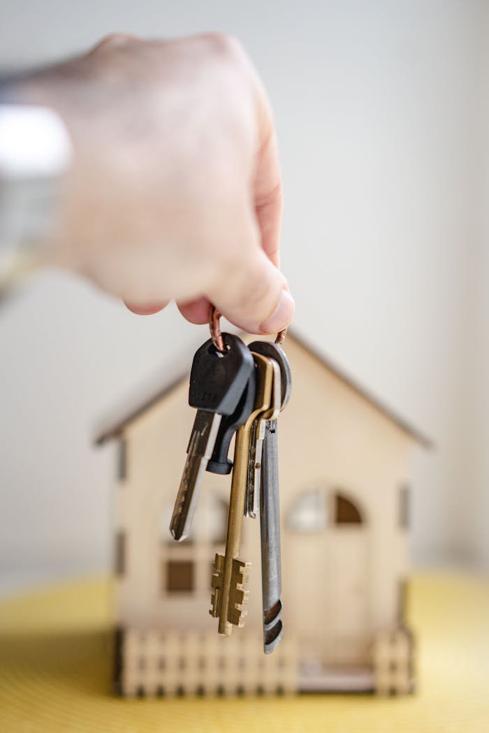 heros-img Close-up of a hand holding keys with a miniature wooden house in the background, symbolizing real estate investment.